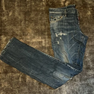 Blå distressed jeans från Dsquared2 - Säljer ett par blå jeans från Dsquared2 med slitna detaljer och snyggt tvättad look. Klassisk femficksmodell med normal passform och raka ben. Jeansen har coola slitningar och en röd Dsquared2-tag vid gylfen. Perfekta för en avslappnad streetstil.