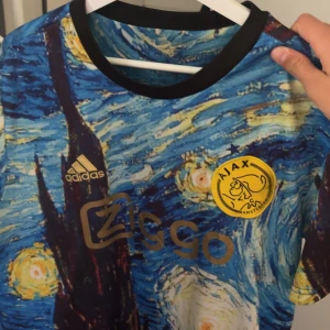 Ajax x Adidas Van Gogh fotbollströja - Unik Ajax fotbollströja från Adidas med Van Gogh-inspirerat mönster i blått, gult och svart. Tröjan har korta ärmar, svart krage och Ajax-logga i gult på bröstet. Ziggo sponsortryck i guld. Perfekt för dig som vill sticka ut på läktaren eller planen.