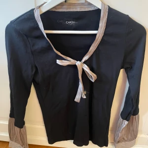 Svart topp med knyt och volangärm - Supersnygg svart topp från Caroll med långa ärmar och unika randiga detaljer i beige vid ärmslut och halsringning. Knytband framtill ger en extra touch. 