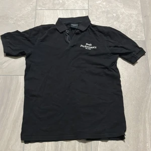 Svart Peak Performance pikétröja - Klassisk svart pikétröja från Peak Performance med broderad logga på bröstet. Tröjan har krage, knappar vid halsen och korta ärmar. Tillverkad i bomull för en skön och avslappnad känsla.