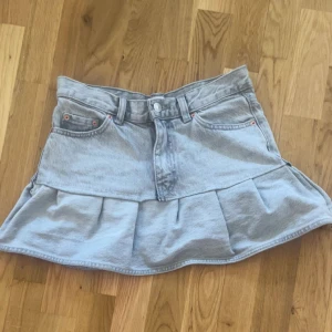 Ljusgrå jeanskjol med volang - Snygg ljusgrå jeanskjol med volangkant och klassiska fickor fram och bak. Kjolen har bälteshällor, knapp och dragkedja framtill. Perfekt för dig som gillar en cool och avslappnad stil.