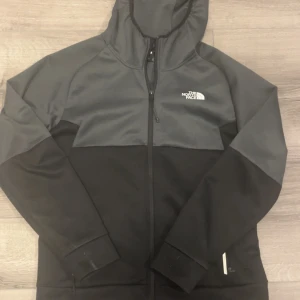 Svart och grå vindjacka från The North Face - Snygg svart och grå vindjacka från The North Face med huva och hel dragkedja. Jackan har logga på bröstet och ryggen, samt praktiska fickor. Perfekt för dig som gillar streetwear och vill ha en stilren look.
