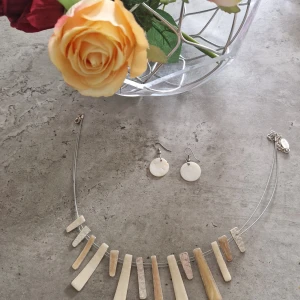 Set med halsband och örhängen i ben - Unikt smyckeset med halsband och matchande örhängen i benfärgad och vit nyans. Halsbandet har flera avlånga benliknande hängen på tunn metalltråd och örhängena är runda, släta och vita. Perfekt för dig som gillar naturliga material och stilren design.