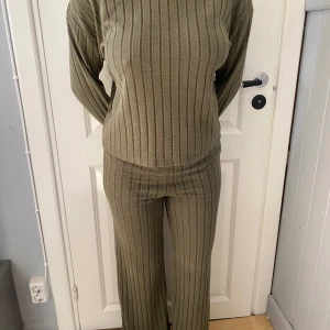 Olivgrön set ribbad  i mjukt tyg - Tvådelad olivgrön set med långärmad topp och byxa. Setet är ribbat med vertikala ränder och har en relaxed passform. Materialet känns mjukt och stretchigt, perfekt för en chill och snygg look. Byxorna är raka och toppen har en enkel, klassisk skärning.