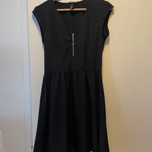 Svart kortklänning från H&M stl 36 - Svart kortklänning från H&M med v-ringning och en snygg dragkedjedetalj framtill. Klänningen har korta ärmar och markerad midja med lätt utställd kjol. 