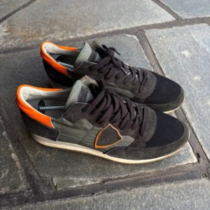 Svarta sneakers med orange detalj - Snygga sneakers från Philippe Model i svart mocka och textil med gråa paneler och orange detaljer vid häl och logga. Skorna har snörning och en vit platt sula som ger en sportig vibe. Perfekta för dig som gillar streetstyle och unika färgkombinationer.