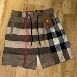 Rutig Burberry shorts med snörning - Snygga shorts från Burberry i klassiskt rutigt mönster med beige, svart, vit, brun och röd. De har elastisk midja med svart snörning och sidofickor. Perfekta för sommaren och ger en lyxig vibe till din outfit.
