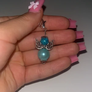 Blå ängel med pärldetalj - Unik lyckoängel format  som en ängel med silverfärgade vingar, en glittrig blå pärla upptill och en större ljusblå pärla nedtill. Fästet är i silverfärg och örhänget har en söt och lekfull design som sticker ut.