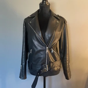 Svart skinnjacka från Missguided - Säljer en svart skinnjacka från Missguided. Jackan har silvriga dragkedjor, bred krage, axelklaffar och bälte i midjan. Långärmad modell med dragkedjor vid ärmsluten och flera fickor framtill. Sitter väldigt fint på.