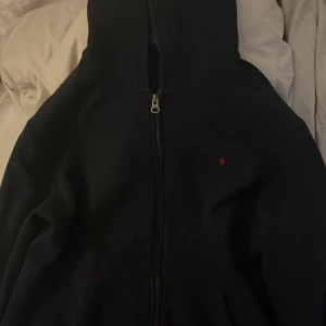 Mörkblå hoodie från Polo Ralph Lauren  - Mörkblå Hoodie från Polo Ralph Lauren i storlek XL. Klassisk design med dragkedja framtill, huva och rött broderat Polo-logo på bröstet. Tillverkad i mjuk bomull, perfekt för chill dagar. Snygg och enkel att matcha med allt.