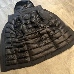 Svart dunjacka med huva från UBR - Säljer en svart dunjacka från UBR med lång passform och huva. Jackan har stilren design, diskreta fickor och quiltad insida. Perfekt för kalla dagar när du vill hålla värmen och ändå se clean ut.