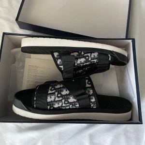 Svarta och vita sandaler från Dior - Snygga sandaler från Dior med svart och vitt mönster, breda remmar med justerbara spännen och tydlig Dior-logga. Ovandelen är i textil med grafiskt mönster och sulan är vit och svart för en clean kontrast. Perfekta för en trendig och avslappnad stil. 