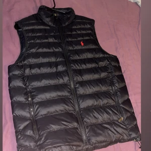 Svart dunväst från Polo Ralph Lauren - Snygg svart dunväst från Polo Ralph Lauren med klassisk röd logga på bröstet. Västen har horisontella quiltade paneler, hög krage och dragkedja framtill samt två praktiska fickor med dragkedja. Perfekt för lager-på-lager och stilren look. Nästan oanvänd.