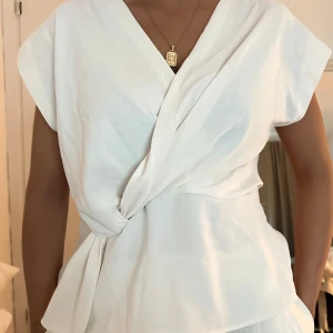 zara vit blus med knytning - Säljer en stilren vit omlottopp med knyt i midjan och v-ringad hals. Toppen har korta ärmar och en avslappnad passform. Perfekt för dig som gillar minimalistisk och fräsch stil. Materialet känns mjukt och svalt mot huden.