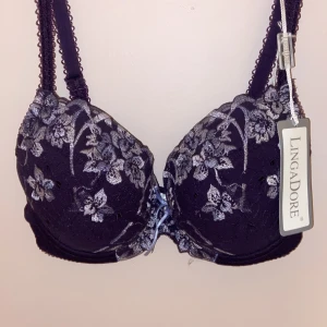 Lila blommig bh från LingaDore 80C - Helt ny.      Superfin lila bh från LingaDore med blommigt spetsmönster i silverton. Dekorerad med en söt rosett och liten glittrig sten mellan kuporna. Kuporna är vadderade och axelbanden har fina spetsdetaljer. Perfekt för dig som gillar snygga detaljer och blommigt.