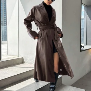 Brun skinn trenchcoat - Snygg lång brun trenchcoat i skinnimitation med bälte i midjan och breda slag. Jackan har långa ärmar med knäppning vid ärmslut och en stilren, minimalistisk look. Perfekt för dig som vill sticka ut med en trendig och edgy vibe.