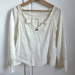 Vit blus med spets och rosett - Supersöt vit blus från Odd Molly med broderat mönster, v-ringning och liten rosett framtill. Blusen har långa ärmar med spetsdetaljer och en lös, avslappnad passform. Perfekt för dig som gillar romantisk och bohemisk stil. 💗