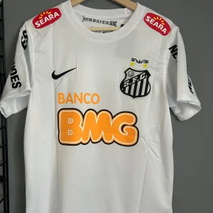 Vit Santos Neymar Jr fotbollströja Nike - Santos FC matchtröja i vitt med Neymar Jr och nummer 11 på ryggen. Tröjan har svarta och orangea tryck, klubbmärke på bröstet och flera sponsorer. Klassisk rund hals och korta ärmar. Tillverkad i lätt och ventilerande material, perfekt för fotbollsälskare. Helt ny med tagg en replika 