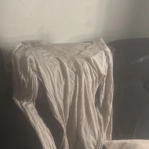 Beige långärmad topp med spetsdetaljer - Supersöt beige långärmad topp med v-ringning och fina spetsdetaljer både vid halsringningen och ärmsluten. Tunt och mjukt material som känns skönt mot huden. Perfekt för dig som gillar romantiska och feminina vibes.