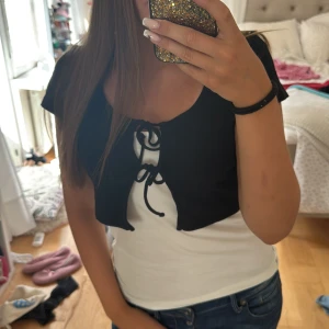 Svart bolerotopp  - Svart kortärmad bolerotopp från H&M, den har en croppad passform och snygg snörning framtill. Tunt och mjukt bomullsmaterial som sitter skönt och ger en trendig layering-effekt över linnen eller klänningar. Perfekt för att lyfta din outfit med en cool detalj!