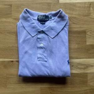 Ralph Lauren piké - Säljer en ljuslila pikétröja från Polo Ralph Lauren i bomull. Klassisk piké med krage, två knappar och broderad logga på bröstet. Kortärmad och normal passform, perfekt för vår och sommar. Snygg och stilren färg som sticker ut utan att bli för mycket. Pikén har några skavanker som man ser på de sista två bilderna och därav priset ;)