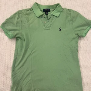 Grön pikétröja från Ralph Lauren - Snygg ljusgrön pikétröja från Polo Ralph Lauren med klassisk krage, korta ärmar och broderad logga på bröstet. Tröjan har knappar vid halsen och är tillverkad i bomull för en skön och luftig känsla.