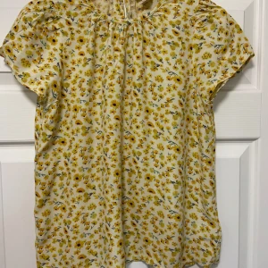 Blommig blus från Holly & Whyte S - Knappt använd! Supersöt blommig blus från Holly & Whyte by Lindex i storlek S. Topp i ljusgul nyans med små gula och lila blommor, rund halsringning och korta ärmar. Knäppning med guldfärgad knapp i nacken. Perfekt för vår och sommar! I ltunt luftigt viskos tyg för fint fall.