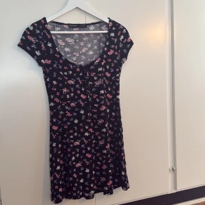 Blommig kortklänning från Bershka - Supersöt svart kortklänning från Bershka med små blommor i rosa, lila och vitt. Klänningen har korta ärmar, rundad urringning och en lätt utställd passform. Klänningen har även en liten knytning framtill. Skulle säga att klänningen är kort även om det är storlek S. Perfekt för sommarens alla tillfällen!