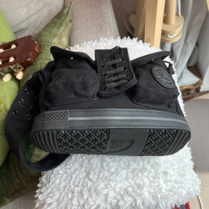 Svart Converse axelväska med snörning - Cool svart axelväska från Converse i canvas med tydlig sneakers-inspirerad design. Väskan har snörningsdetalj framtill, två fickor och klassisk Converse-logga på sidan. Botten är i gummi med mönster som på en sneakersula.