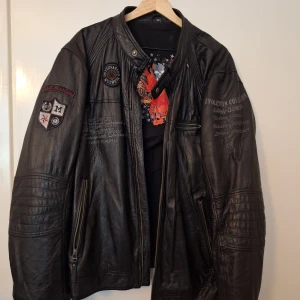 Svart skinnjacka Zeda med patches - Riktigt snygg svart skinnjacka från Zeda, designad av Roberto Bertolucci. Jackan har coola broderade patches och detaljer på ärmarna, dragkedjor och tryck på bröstet. Tillverkad i äkta skinn med polyesterfoder. Perfekt för dig som vill sticka ut med en edgy stil.