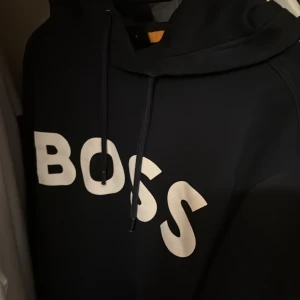 Mörkblå hoodie från BOSS med tryck - Snygg mörkblå hoodie från BOSS med stor vit logga framtill. Klassisk modell med huva och dragsko, samt känguruficka. Perfekt för dig som gillar stilrena och bekväma plagg med streetkänsla.