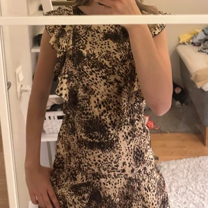 Leopardmönstrad kortklänning AX Paris - Snygg kortklänning från AX Paris med leopardmönster i beige och svart. Klänningen har volangdetaljer vid axlar och nederkant, är ärmlös och har en lätt utsvängd form. Perfekt för dig som vill sticka ut med ett djurmönster och coola detaljer.