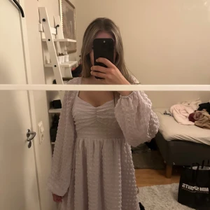 Ljuslila kortklänning från NA-KD - Supersöt ljuslila kortklänning från NA-KD med puffiga långärmar och fyrkantig ringning. Klänningen har ett strukturerat tyg med rutigt mönster och rynk vid bysten. Perfekt för dig som gillar romantisk och trendig stil.