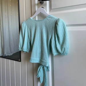 Mintgrön omlottopp med puffärm - Supersöt mintgrön topp med puffiga korta ärmar och knyt i ryggen. Toppen har en strukturerad, krinklad yta och v-ringnad baksida. Perfekt för dig som gillar trendiga detaljer och en feminin look.