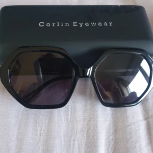 Svarta solglasögon från Corlin Eyewear - Snygga svarta solglasögon från Corlin Eyewear med stora, åttakantiga bågar i blank plast. Glasen är mörka och ger en cool statement-look. Perfekta för dig som vill sticka ut och addera lite edge till din stil.