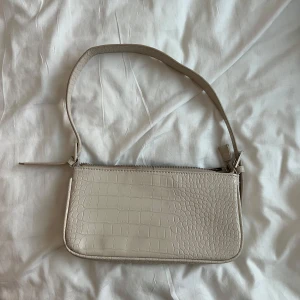 Beige axelväska från Gina Tricot - Snygg beige axelväska från Gina Tricot med croco-mönstrad yta och justerbart axelband. Väskan har dragkedja upptill och är perfekt för att bära det viktigaste. Stilren och trendig design som lyfter vilken outfit som helst.
