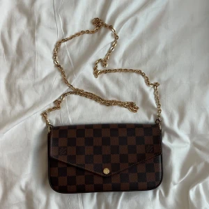 Brun kuvert axelväska  - Säljer en axelväska  med bruna och mörkbruna rutor. Väskan har guldfärgad kedja, röd insida och medföljande plånbok och korthållare. Perfekt storlek för det viktigaste och riktigt snygg detalj till outfiten.