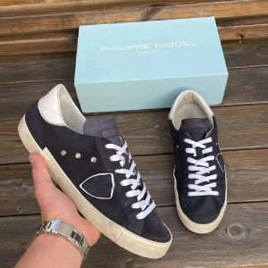 Philippe Model sneakers - Snygga Philippe Model skor i mocka med vita detaljer och vit sula. Skorna har vita skosnören, silverfärgade nitar och en ikonisk sköld på sidan. Hälpartiet är i vitt skinn och sulan är platt och beige. Perfekta för en stilren och trendig look.