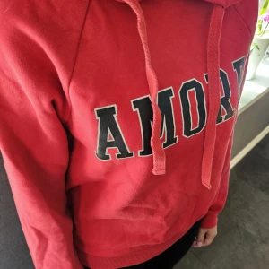 Röd hoodie med AMORE från Gina Tricot - Röd hoodie från Gina Tricot med svartvitt AMORE-tryck på bröstet. Tröjan har huva med snörning och långa ärmar. Mjuk insida och ribbade muddar vid ärmslut och nederkant. Perfekt för dig som gillar en chill och snygg streetstil. Säljer denna eftersom den inte kommer till användning 