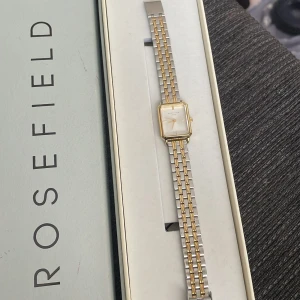 Rosefield rektangulär klocka guld/silver - Stilren klocka från Rosefield med rektangulärt ur i guldton och vit urtavla. Länken är tvåfärgad i silver och guld, vilket ger en snygg kontrast. Klockan har minimalistisk design och metallarmband som känns både klassisk och modern.