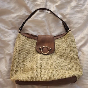 Beige och brun axelväska med gulddetalj - Snygg axelväska i beige flätad look med bruna skinnliknande detaljer och axelrem. Väskan har ett lock med guldfärgad ring framtill, dragkedja på baksidan och blommigt foder inuti. Perfekt storlek för allt du behöver till vardags.