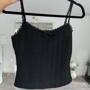Svart ribbad linnetopp med spets - Supersöt svart linnetopp med smala stretchiga axelband och spetskant längs urringningen. Toppen har en liten rosett framtill och ett ribbat mönster som ger en snygg struktur. Perfekt att styla med jeans eller kjol för en trendig look. Blivit en aning nopprig. 💘 Önskar du veta exakta mått? Tveka inte att höra av dig!