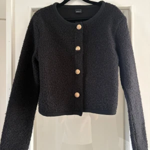 Svart kofta med guldknappar - Svart kofta från Gina Tricot med lång ärm och fyra guldknappar framtill. Koftan är i ett mjukt, stickat material med lite fluffig textur och har en rak, något croppad passform. Perfekt för lager-på-lager och enkel att styla till olika looks.