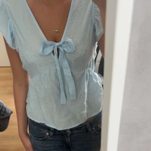 Ljusblå topp VERO MODA - Supersöt ljusblå blus från VERO MODA med v-ringning och knytrosett där framme. Toppen har korta ärm med volang och är tillverkad i ett tunt, luftigt material som känns perfekt nu på sommaren. Tveka inte att fråga om du undrar något! 💌