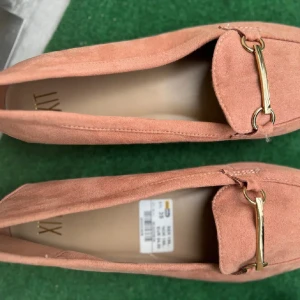 Rosa bellerinaskor från XIT med gulddetalj - Säljer ett par söta ballerina skor i rosa mocka från XIT med guldfärgad metallspänne framtill. Skorna har platt sula och rund tå, vilket gör dem både stilrena och bekväma. Perfekta för sommaren och skolstart, kan stylas med både kjol eller jeans💞