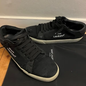 Svarta Saint Laurent sneakers - Svarta sneakers från Saint Laurent med vit broderad logga på sidan. Skorna har lossnat lite på sidan men jag har löst det med superlim de ser värre ut än vad det är. Du får med dustbag och box. Priser är inte hugget // Ian 