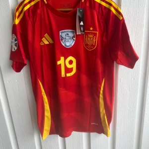 Spanien Lamine Yamal matchtröja Adidas - Säljer en officiell Spanien fotbollströja med Lamine Yamal och nummer 19. Tröjan är röd med gula detaljer och korta ärmar, tillverkad i lätt och ventilerande material. Adidas-logga och landslagsemblem framtill. Matchande blå shorts med gula inslag och landslagsemblem ingår.