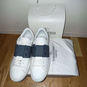 Valentino Garavani Open sneakers vit/blå - Snygga Valentino Garavani Open sneakers i vitt skinn med bred marinblå rem över vristen och vita snören. Klassisk rund tå, platt sula med nitar baktill och diskret guldlogga på plösen. Perfekta för dig som vill ha en clean och lyxig look. Kvitto finns hör av dig vid frågor och funderingar 