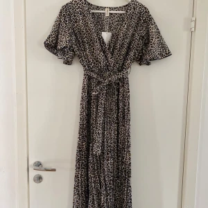 Leopardmönstrad långklänning DEFOX - Snygg långklänning från DEFOX med leopardmönster i svart, brun och beige. Klänningen har omlottliv, v-ringning, korta vida fjärilsärmar, plisserad kjol och knytskärp i midjan. Perfekt för dig som vill sticka ut med ett djurmönster och flowig passform. Ny med prislappen kvar. 
