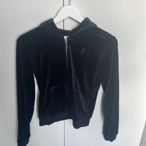 Mörkblå velour hoodie Juicy Couture - Mjuk och lyxig mörkblå hoodie från Juicy Couture i glansig velour. Tröjan har dragkedja med J-detalj, broderad logga på bröstet, huva och känguruficka. Perfekt för en chill och trendig look.